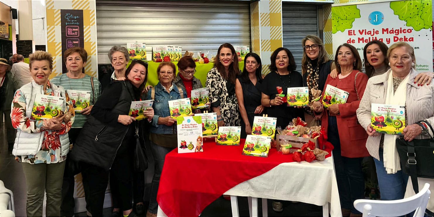 Grupo de mujeres sonrientes sosteniendo libros del proyecto "El Viaje Mágico de Melita y Deka" en un evento comunitario. Fondo con decoración colorida y mesas con productos. Ideal para promover la lectura y la participación de la comunidad.